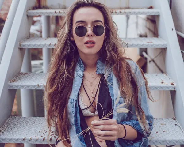 Frau mit langen Haaren und Sonnenbrille sitzt auf einer Metalltreppe. Sie trägt ein offenes Jeanshemd, das sie lässig mit einem Spitzen-Bralette kombiniert.