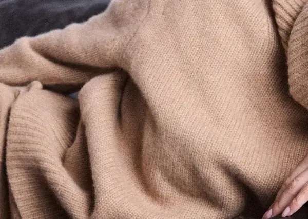 Eine Frau in einem beigen Kaschmirpullover liegt entspannt auf einem dunklen Sofa. Das Kaschmir waschen ist wichtig für die Pflege.