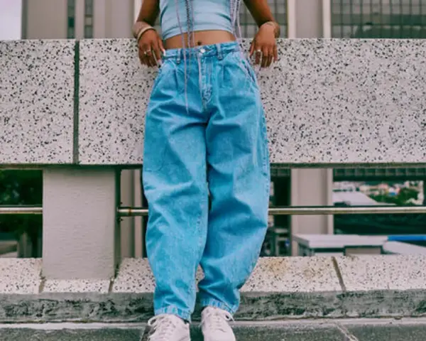 Person in einem lässigen baggy jeans outfit mit Crop-Top und weißen Sneakern steht auf einer Betonmauer.
