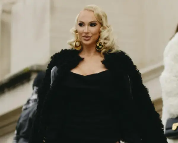 Eine Frau im Stil einer "mob wife" mit blonden Locken, großen goldenen Ohrringen und einem schwarzen Pelzmantel.
