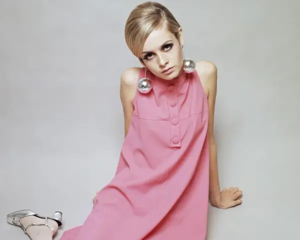 Ein Model trägt ein rosa Kleid mit großen Kugell-Ohrringen, typisch 60er Jahre Kleidung. Silberne Schuhe runden den Look ab.