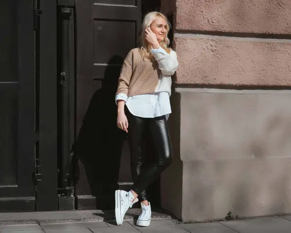 Eine Frau kombiniert elegant schwarze Hose mit einem zweifarbigen Pullover und weißem Hemd. Weiße Plateau-Sneaker runden den Look ab.
