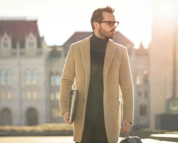 Ein Mann im old money style, mit Brille und beigem Mantel, trägt einen Laptop und eine Reisetasche.