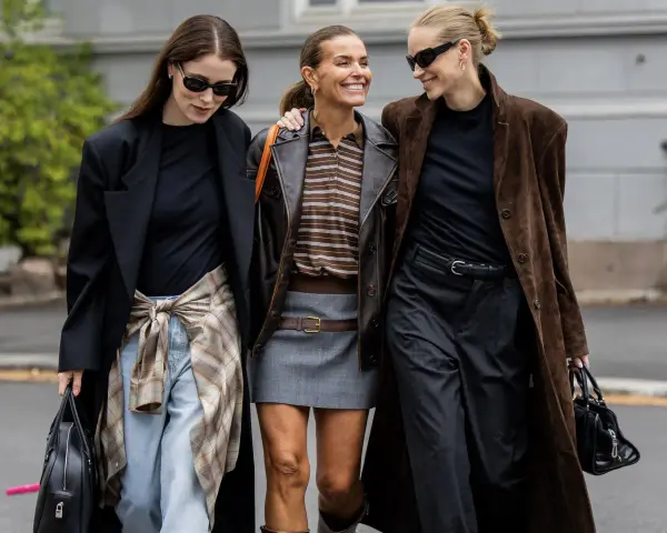 Drei Frauen in komischer Mode, die lachend nebeneinander gehen. Eine trägt einen braunen Lederblazer, die andere einen langen braunen Mantel.