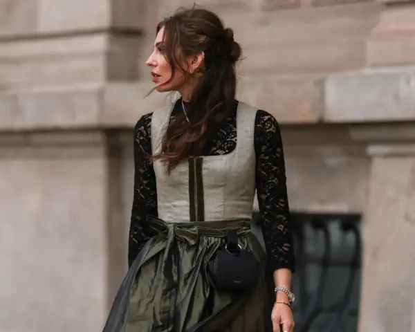 Eine Frau trägt eine graue Weste über einer schwarzen Spitzenbluse. Sie kombiniert die schwarze Bluse mit einem langen, olivgrünen Rock und einer kleinen schwarzen Tasche.