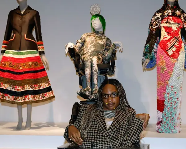 Bei einer Vernissage werden extravagante Kleider auf Schaufensterpuppen präsentiert. Eine Frau im Rollstuhl sitzt davor.