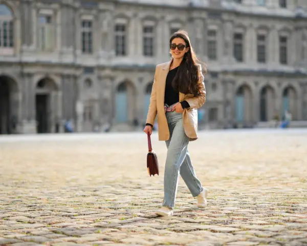 Frau in weiter Jeans und blauem Hemd mit goldener Clutch und Glitzer-Schuhen überquert Straße.