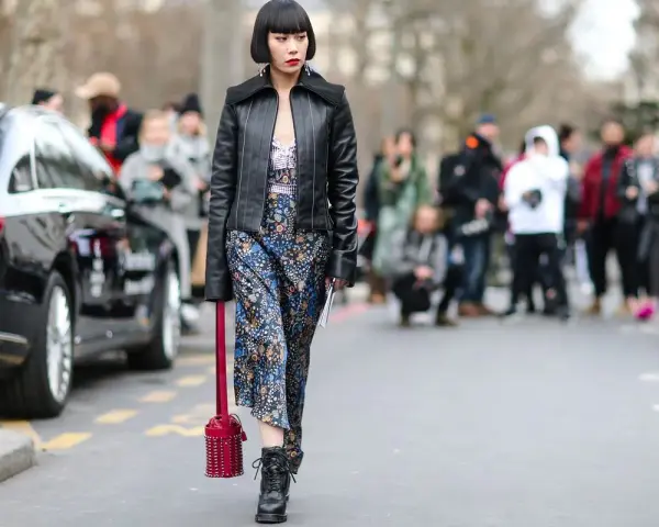 Frau im typisch 90er Grunge Style mit Lederjacke, geblümtem Kleid und Stiefeln. Ein roter Nietenbeutel rundet den Look ab.