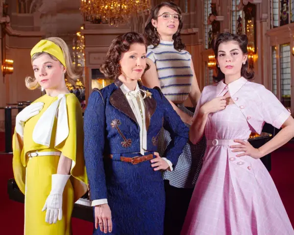 Vier Frauen in stilvollen Outfits, die an die Kleiderordnung des Theater des Westens erinnern, posieren vor einem opulenten Hintergrund.