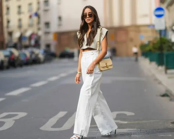 Stilvolle Dame in Paris, die eine weiße Leinenhose kombiniert mit einem kurzen Top und einer Umhängetasche trägt.