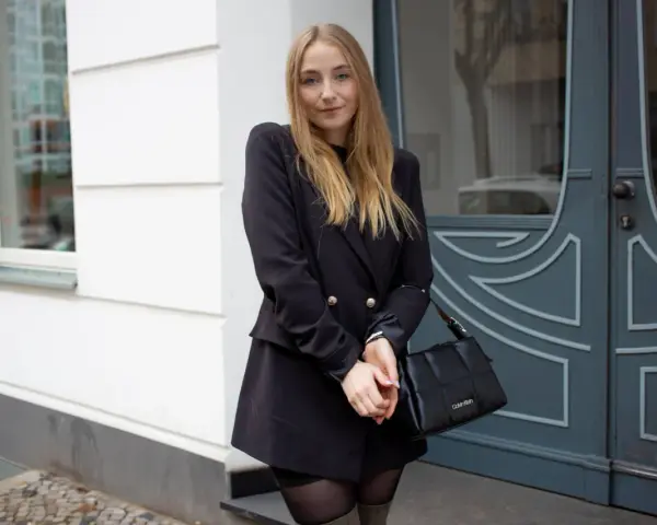 Frau mit blonden Haaren trägt ein schwarzes Kleid mit Blazer, kombiniert mit einer schwarzen Tasche.