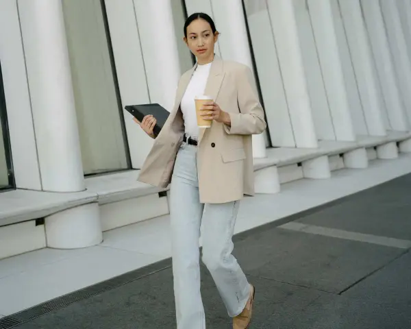 Frau im smart casual Dresscode mit Blazer, Jeans und Kaffee. Sie hält ein Tablet.