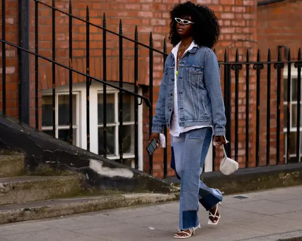 Frau in Jeans-Patchwork-Hose und Jeansjacke, ein Trend für den Jeans-Trend 2026. Weiße Sonnenbrille, weiße Sandalen.