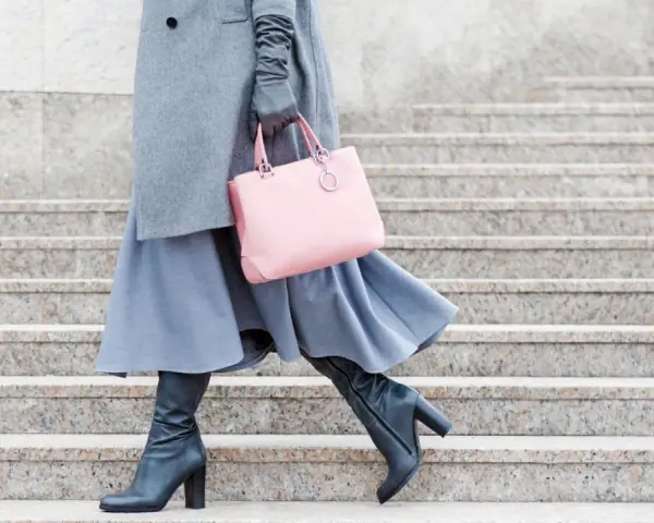Stilvoll **Kleider im Winter tragen**: Grauer Mantel, blauer Rock, schwarze Stiefel und eine rosafarbene Tasche auf Steinstufen.