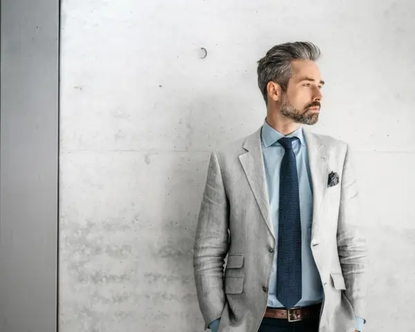 Stilvolles herren outfit sportlich elegant ab 50: Mann mit grauem Sakko, blauem Hemd und Krawatte vor Betonwand.