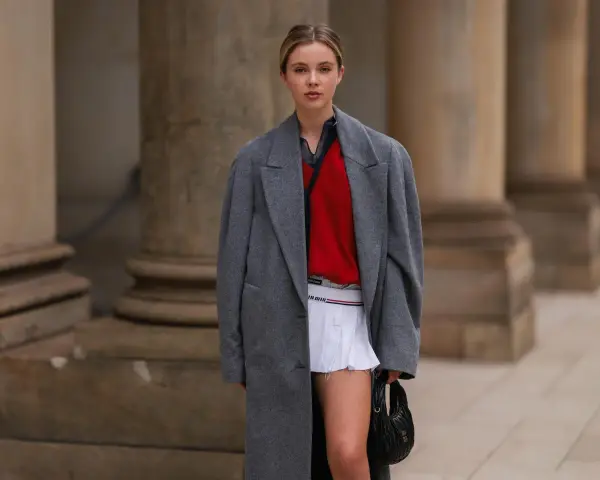 Ein junges Model präsentiert einen preppy look mit grauem Mantel, rotem Pullover und weißem Faltenrock.
