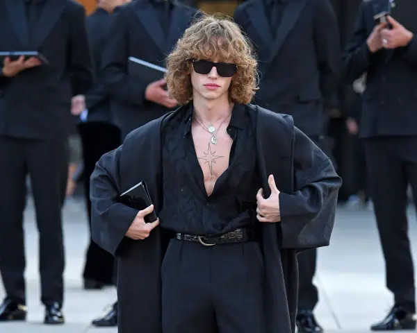 Ein Mann mit lockigem Haar und Sonnenbrille trägt eine schwarze, offen geknöpfte Bluse, weite Hosen und einen Mantel. Die Kleidung wirkt typisch 80er Jahre.