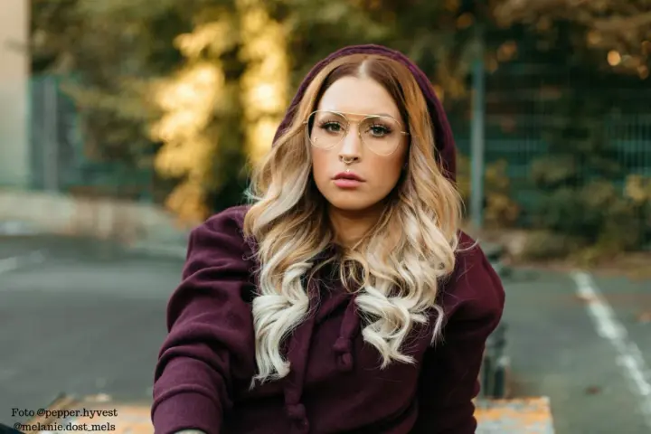 Frau mit blonden Haaren und Brille trägt einen Kapuzenpullover in dunklem Beere. Die Farbe Beere kombiniert sie gekonnt mit ihrem Outfit.
