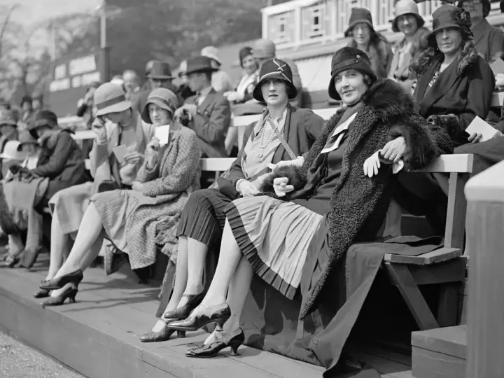 Frauen in eleganten Hüten und Kleidern, ein Bild der Weimarer Republik Mode, sitzen auf einer Tribüne.