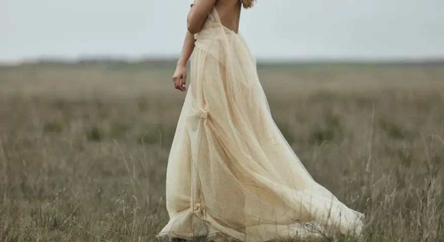 Braut in einem fließenden, cremefarbenen Kleid mit Tupfenmuster, das perfekt zum **dresscode boho hochzeit** passt. Sie steht in einer weiten, grasbewachsenen Ebene.