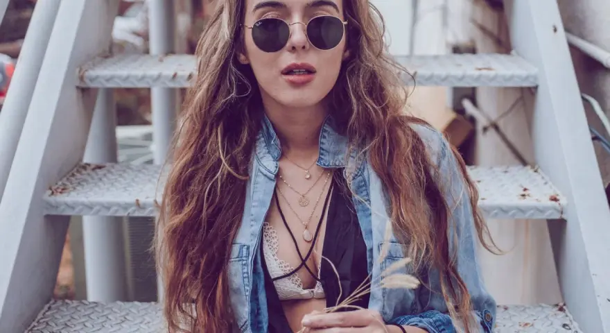 Frau mit langen Haaren und Sonnenbrille sitzt auf einer Metalltreppe. Sie trägt ein offenes Jeanshemd, das sie lässig mit einem Spitzen-Bralette kombiniert.
