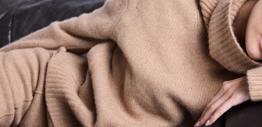 Eine Frau in einem beigen Kaschmirpullover liegt entspannt auf einem dunklen Sofa. Das Kaschmir waschen ist wichtig für die Pflege.