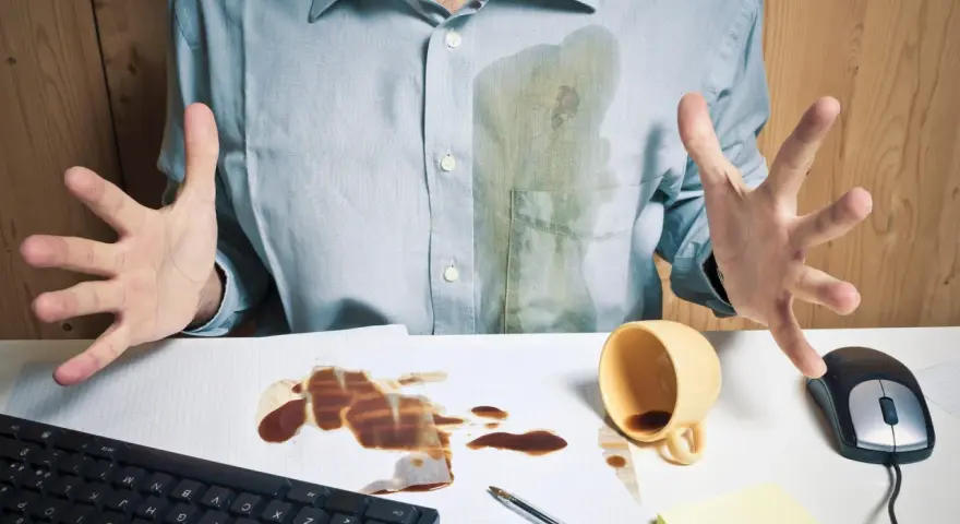 Mann mit verschüttetem Kaffee auf Hemd und Schreibtisch. Eingetrocknete Kaffeeflecken entfernen ist eine Herausforderung.