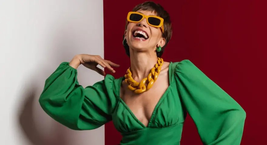 Lachende Frau in grünem Kleid mit auffälligem gelben Statement-Collier und Sonnenbrille, die aktuelle Trendfarben verkörpert.