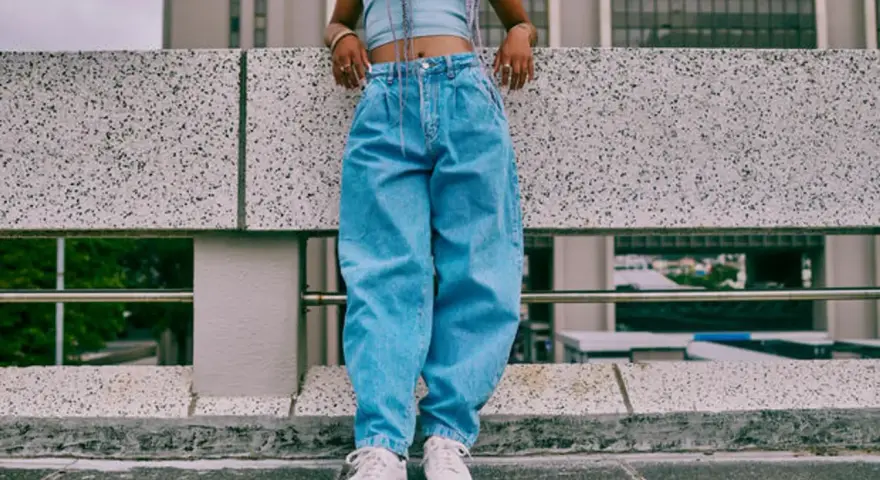 Person in einem lässigen baggy jeans outfit mit Crop-Top und weißen Sneakern steht auf einer Betonmauer.