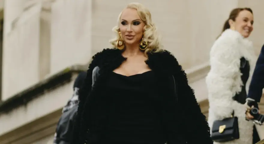 Eine Frau im Stil einer "mob wife" mit blonden Locken, großen goldenen Ohrringen und einem schwarzen Pelzmantel.