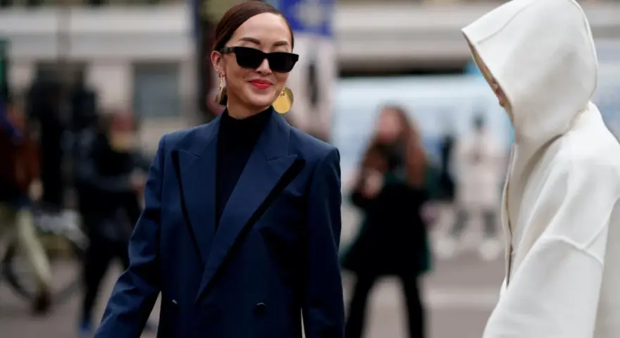 Eine Frau mit Sonnenbrille und auffälligem Schmuck trägt einen dunkelblauen Blazer über einem schwarzen Rollkragenpullover. Sie kombiniert dunkelblau und schwarz gekonnt.