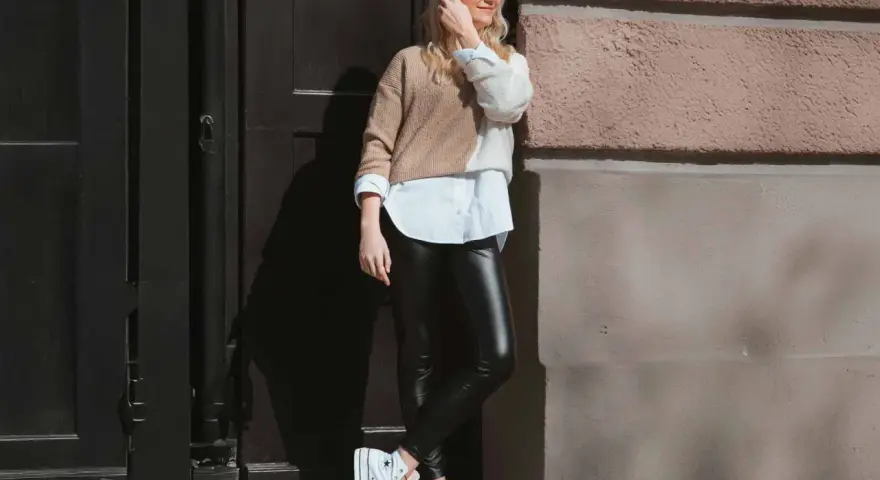 Eine Frau kombiniert elegant schwarze Hose mit einem zweifarbigen Pullover und weißem Hemd. Weiße Plateau-Sneaker runden den Look ab.