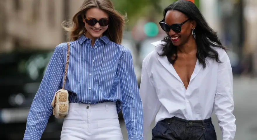 Zwei Frauen in lässiger Kleidung, eine mit blau-weiß gestreifter Bluse und weißer Hose, die andere mit weißer Bluse und dunkler Hose.