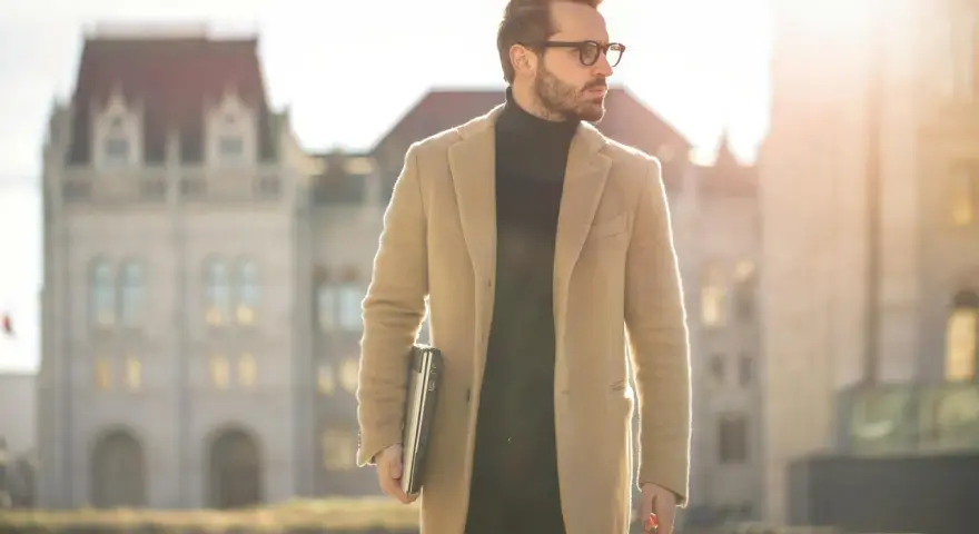 Ein Mann im old money style, mit Brille und beigem Mantel, trägt einen Laptop und eine Reisetasche.