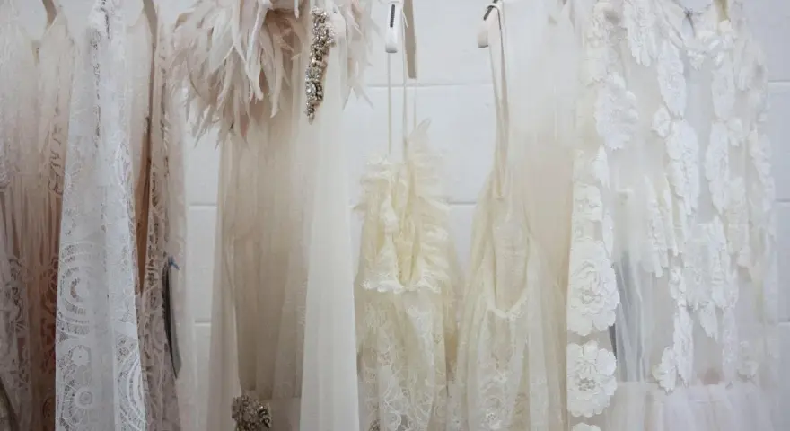 Mehrere zarte, weiße Brautkleider hängen an Kleiderbügeln. Eines hat eine Federverzierung. Die Reinigung eines Hochzeitskleides ist wichtig, damit es schön bleibt.