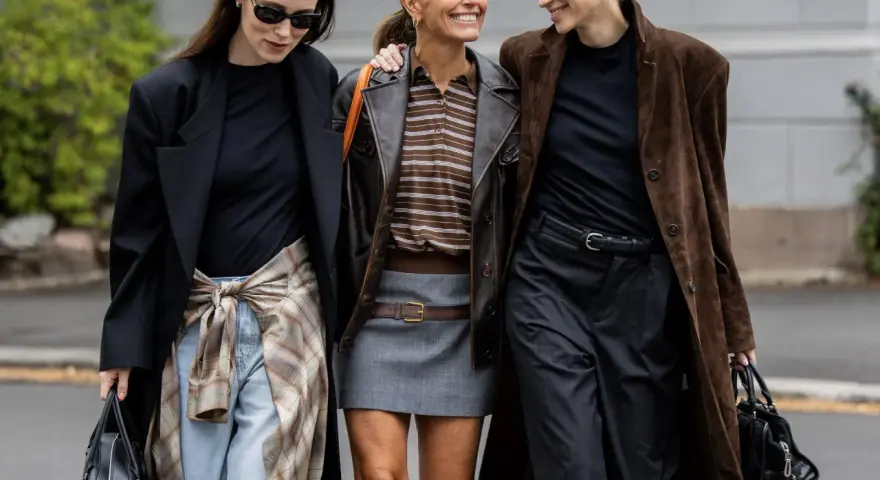Drei Frauen in komischer Mode, die lachend nebeneinander gehen. Eine trägt einen braunen Lederblazer, die andere einen langen braunen Mantel.