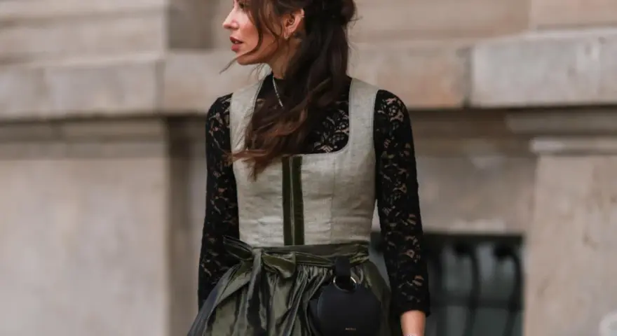 Eine Frau trägt eine graue Weste über einer schwarzen Spitzenbluse. Sie kombiniert die schwarze Bluse mit einem langen, olivgrünen Rock und einer kleinen schwarzen Tasche.