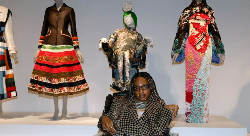Bei einer Vernissage werden extravagante Kleider auf Schaufensterpuppen präsentiert. Eine Frau im Rollstuhl sitzt davor.