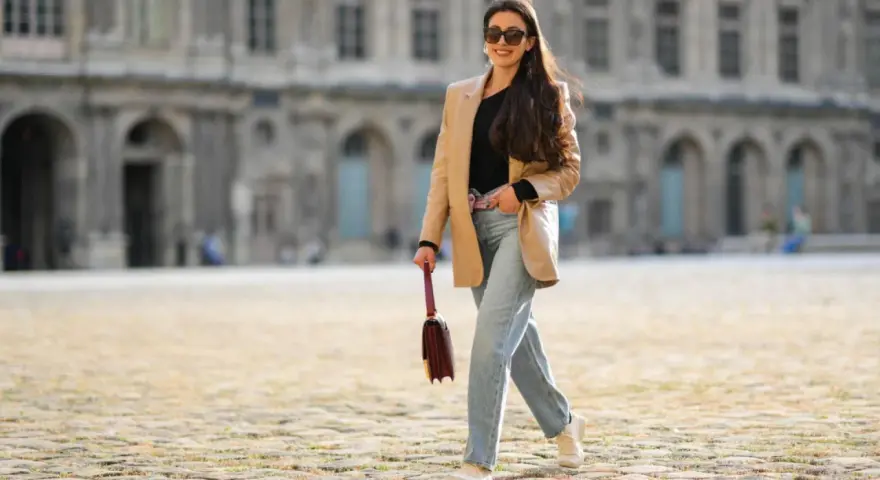 Frau in weiter Jeans und blauem Hemd mit goldener Clutch und Glitzer-Schuhen überquert Straße.