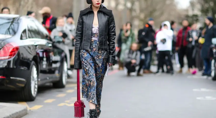 Frau im typisch 90er Grunge Style mit Lederjacke, geblümtem Kleid und Stiefeln. Ein roter Nietenbeutel rundet den Look ab.