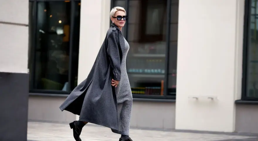Elegante Frau ab 70 mit Kurzhaarschnitt und Sonnenbrille, die in einem langen grauen Mantel und glitzerndem Kleid durch die Stadt geht.