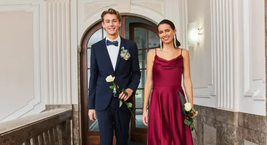 Elegantes Outfit für einen Jugendweihe Gast: Ein junger Mann im dunkelblauen Anzug mit Fliege und eine Frau im weinroten Kleid.