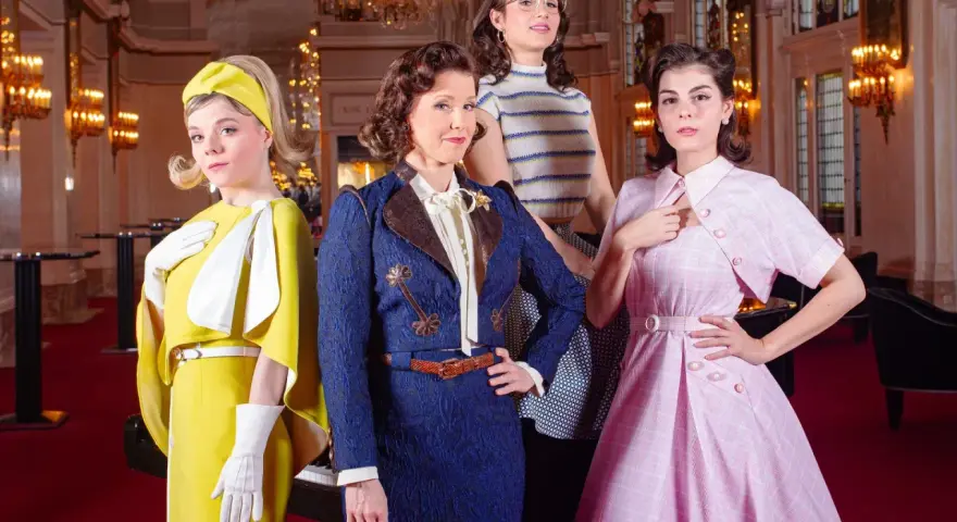 Vier Frauen in stilvollen Outfits, die an die Kleiderordnung des Theater des Westens erinnern, posieren vor einem opulenten Hintergrund.
