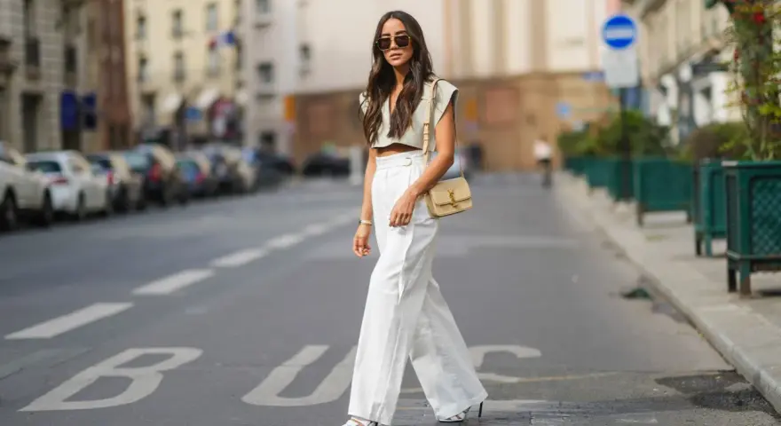 Stilvolle Dame in Paris, die eine weiße Leinenhose kombiniert mit einem kurzen Top und einer Umhängetasche trägt.