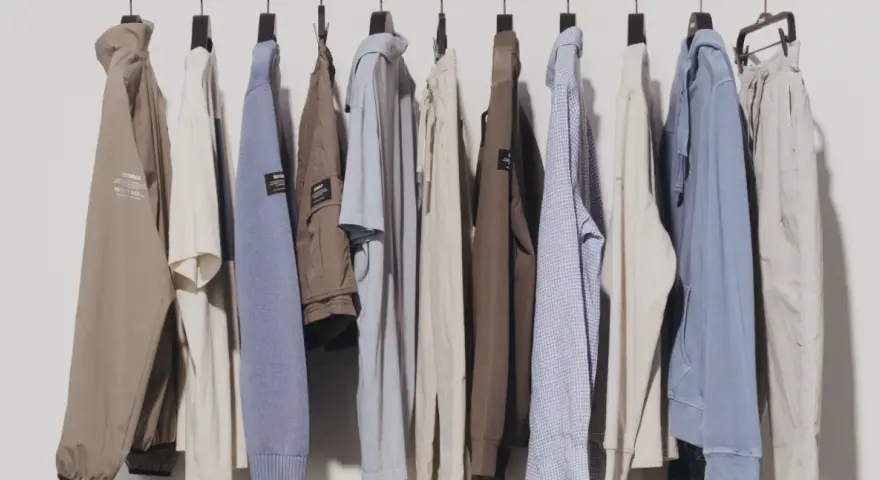 Eine Reihe von Kleidungsstücken, darunter Jacken, Pullover und Hosen, hängen an Kleiderbügeln. Diese zeitlose Kleidung in gedeckten Farben strahlt Komfort und Stil aus.
