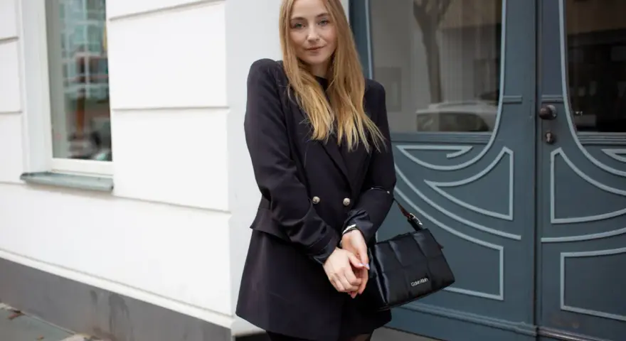 Frau mit blonden Haaren trägt ein schwarzes Kleid mit Blazer, kombiniert mit einer schwarzen Tasche.