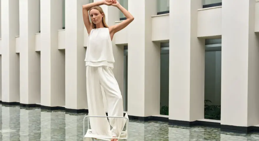 Frau in weißem Outfit mit weiten Palazzo Hosen, elegant gestylt vor moderner Architektur.