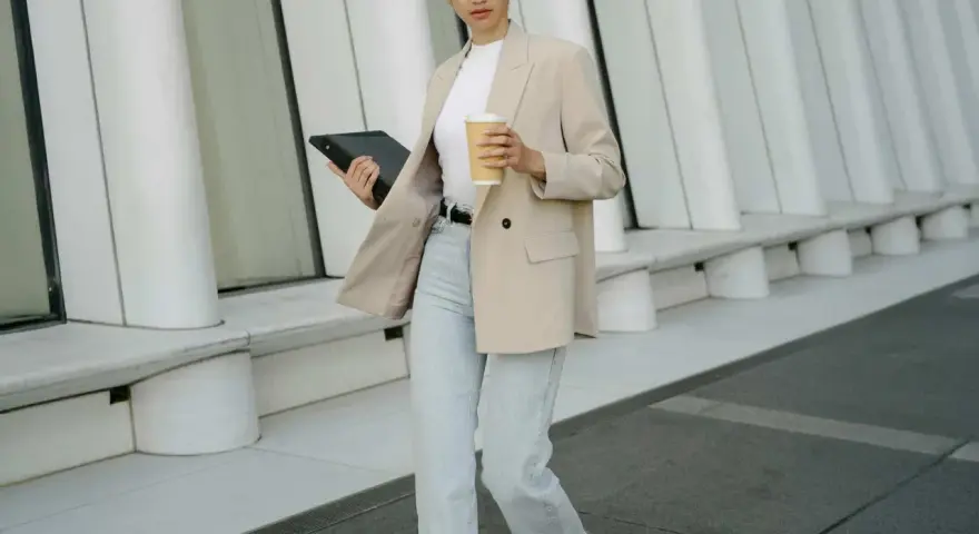 Frau im smart casual Dresscode mit Blazer, Jeans und Kaffee. Sie hält ein Tablet.