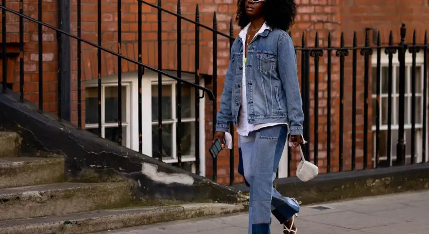 Frau in Jeans-Patchwork-Hose und Jeansjacke, ein Trend für den Jeans-Trend 2026. Weiße Sonnenbrille, weiße Sandalen.