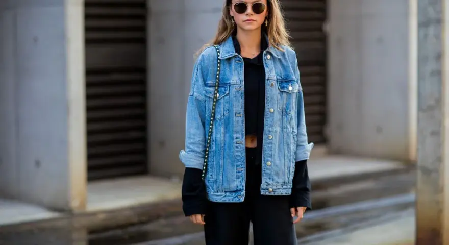 Stylische Frau mit lässiger Jeansjacke kombinieren mit schwarzem Crop-Top und Hose. Sonnenbrille und kleine Tasche runden den Look ab.